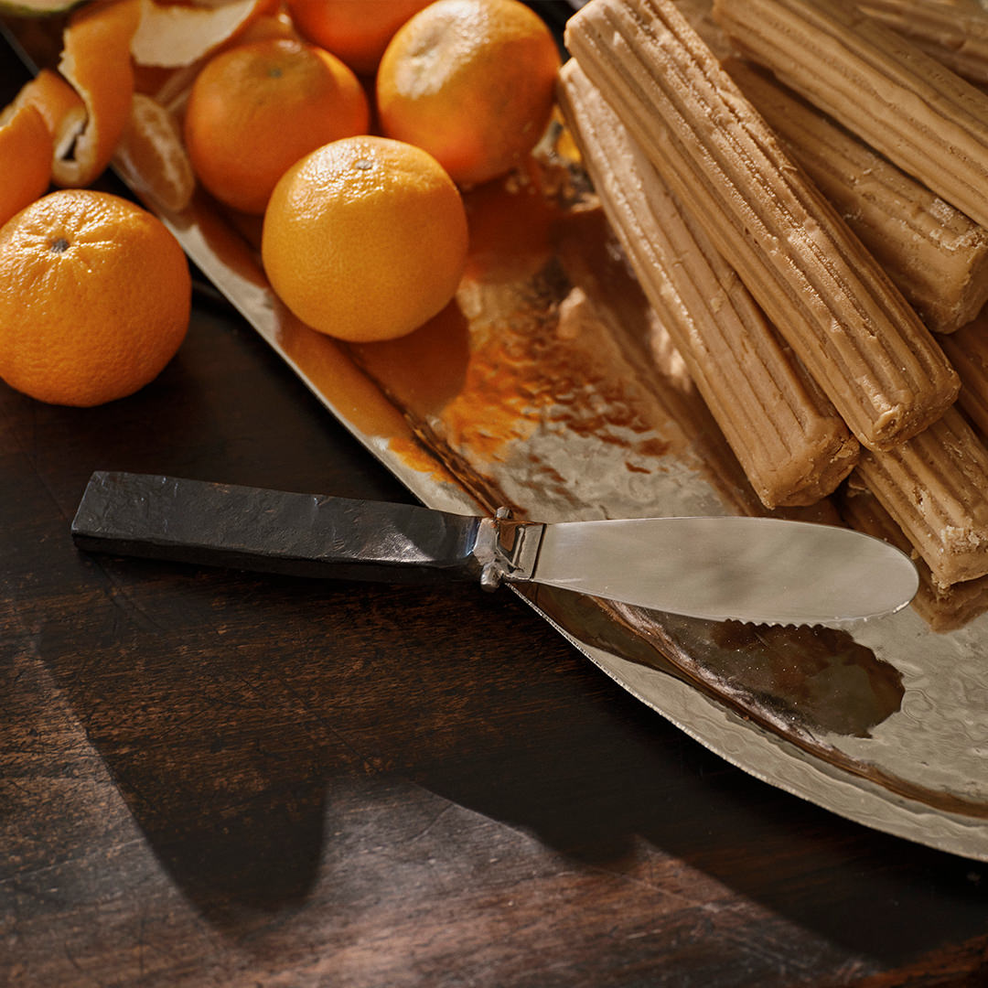 A stainless steel knife with forged handle resting on top of a wood surface and a silver tray. Behind it are some oranges and light brown colored candy sticks.