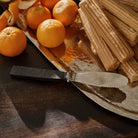 A stainless steel knife with forged handle resting on top of a wood surface and a silver tray. Behind it are some oranges and light brown colored candy sticks.