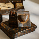 A open iron box with the lid with a heart next to it. Inside the box are some chocolates. The box is on top of another hammered iron box and behind is a box with bread.