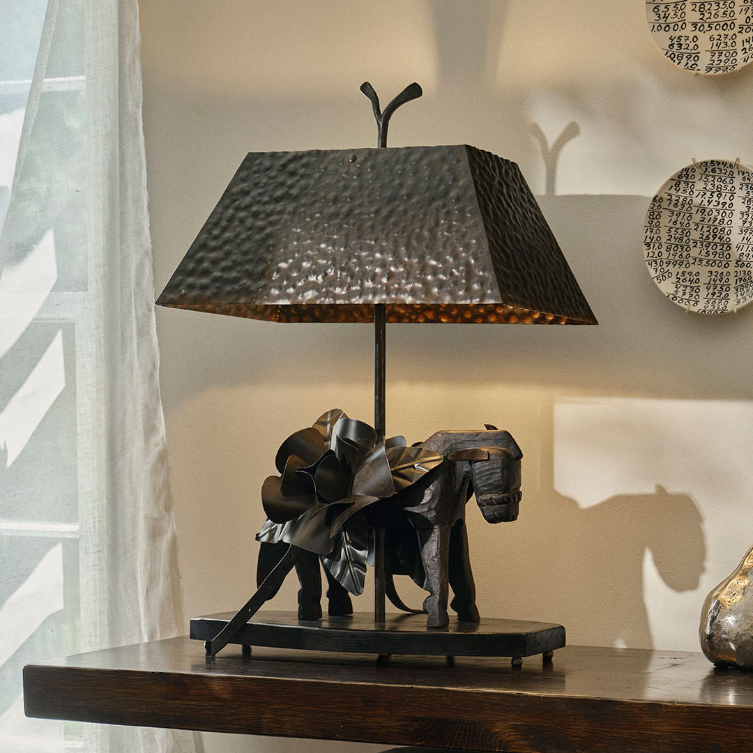 A handcrafted wooden burro with a load of flowers lamp. On top of a wood table and against a white wall and next to a  a curtain.