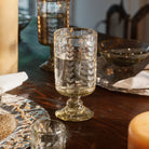 A mouth blown glass engraved with vertical laurel leaves filled with water on top of a wood table. Surrounded by plates, trays and bowls.