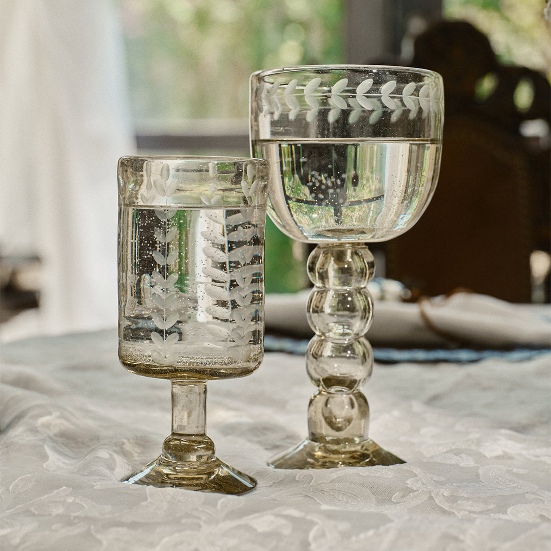 A mouth blown glass engraved with vertical laurel leaves next to a clear glass cup with laurel engravings. Both are filled with water and on top of a withe fabric. On the background is a window.