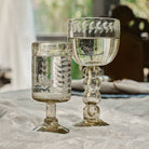 A mouth blown glass engraved with vertical laurel leaves next to a clear glass cup with laurel engravings. Both are filled with water and on top of a withe fabric. On the background is a window.