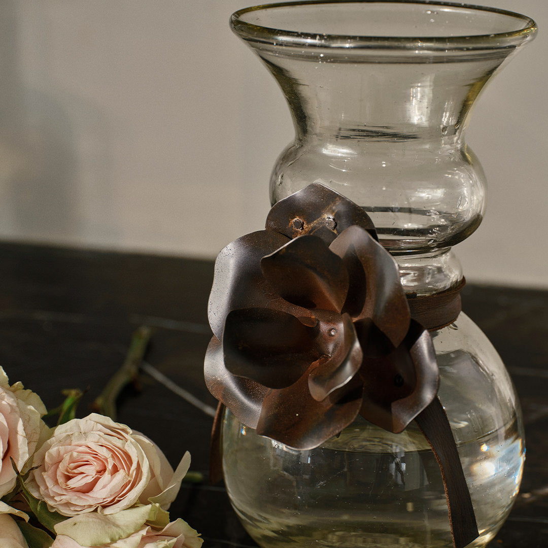 A glass vase with a iron rose and a leather tie. A pink rose to its left.