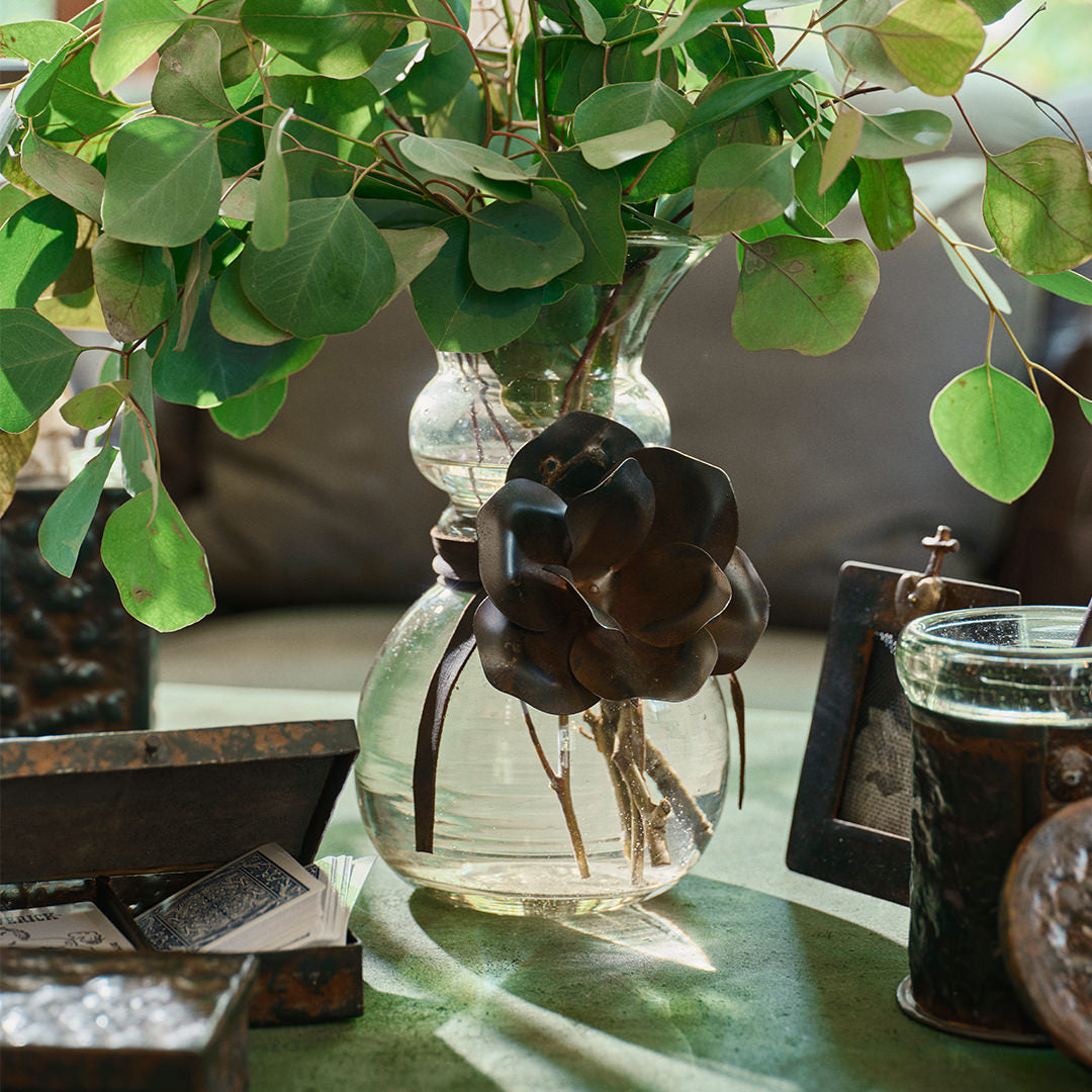 A glass vase with a iron rose and a leather tie.  Inside the vase are green leaves. The vase is surrounded by iron containers and a picture frame.