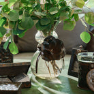 A glass vase with a iron rose and a leather tie.  Inside the vase are green leaves. The vase is surrounded by iron containers and a picture frame.