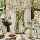 A glass vase with white flowers surrounded by clear glass cups, glasses, candles and small trays with chocolates. Everything is on top of a white fabric. On the  background is a window.