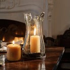 A wood table edge with a glass vase on a iron  vase with a lit yellow candle. next to it is a lit yellow candle. On the background is a white chimney.