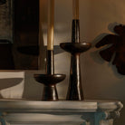 Two  iron primitive and sculptural candlesticks with a candle each. On top of a white surface.