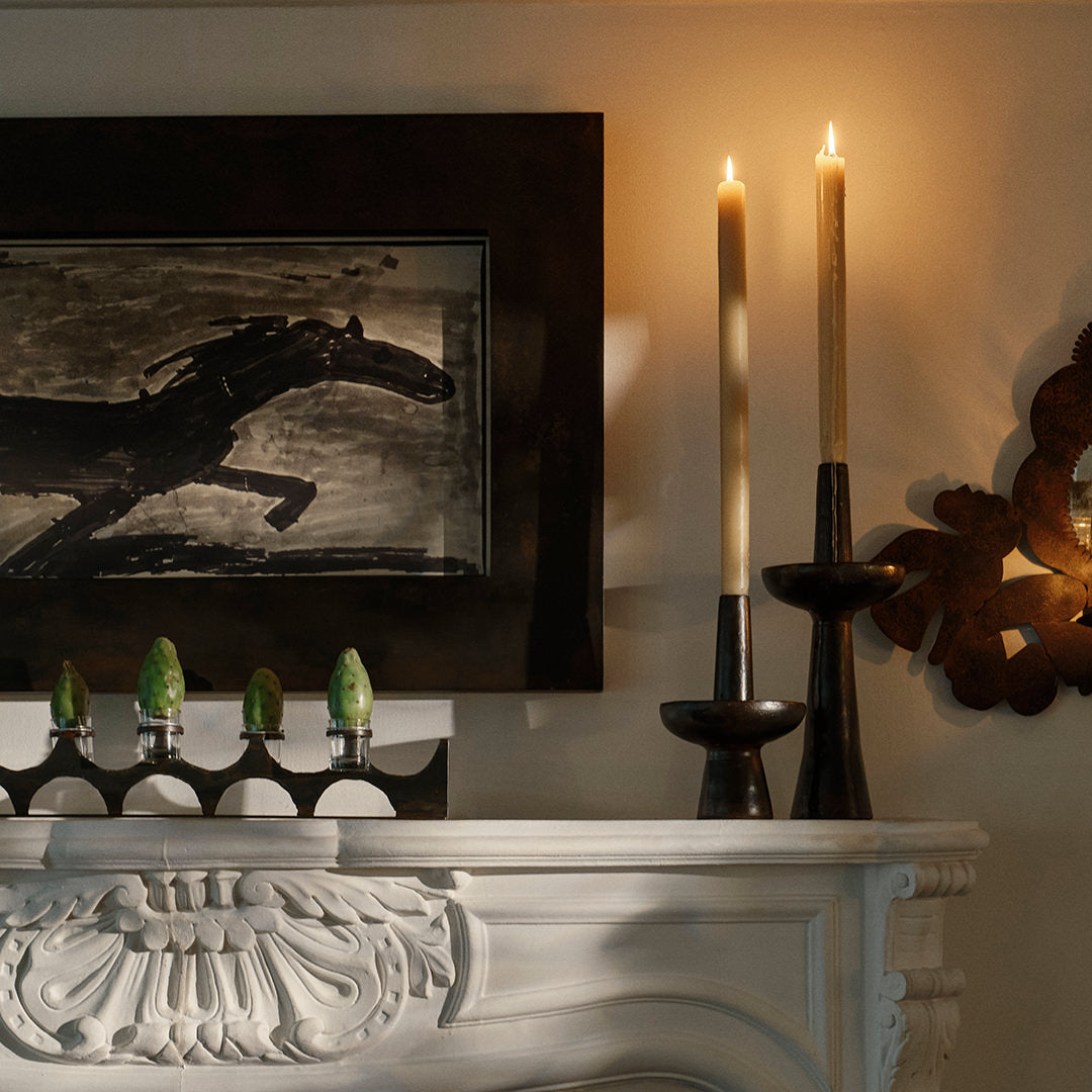 Two  iron primitive and sculptural candlesticks with a candle each. On top of a white surface, Next to a iron sculpture holding green objects and a frame.