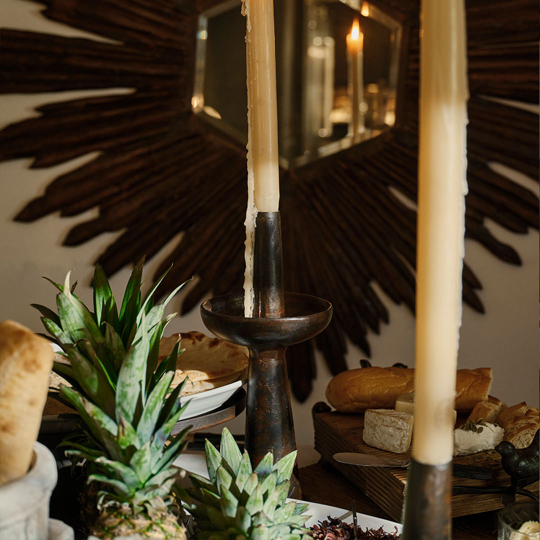 Two  iron primitive and sculptural candlesticks with a candle each. Surrounded by objects such as bread, pineapples, and cheese. On the background is a dark brown mirror frame.