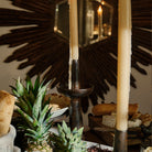 Two  iron primitive and sculptural candlesticks with a candle each. Surrounded by objects such as bread, pineapples, and cheese. On the background is a dark brown mirror frame.