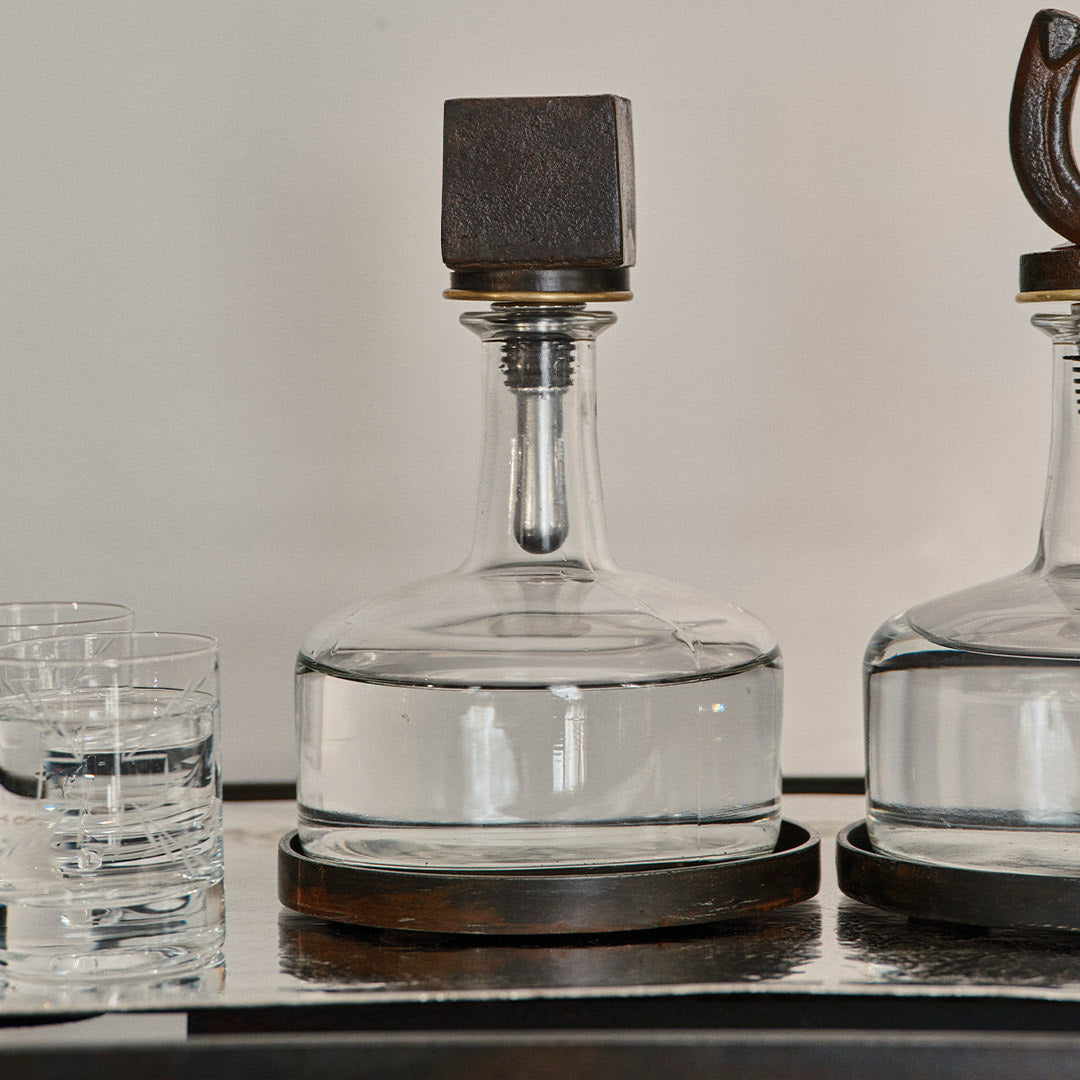 A iron squared top decanter and leather lined iron tray In  the middle of another decanter and two glasses with liquid.
