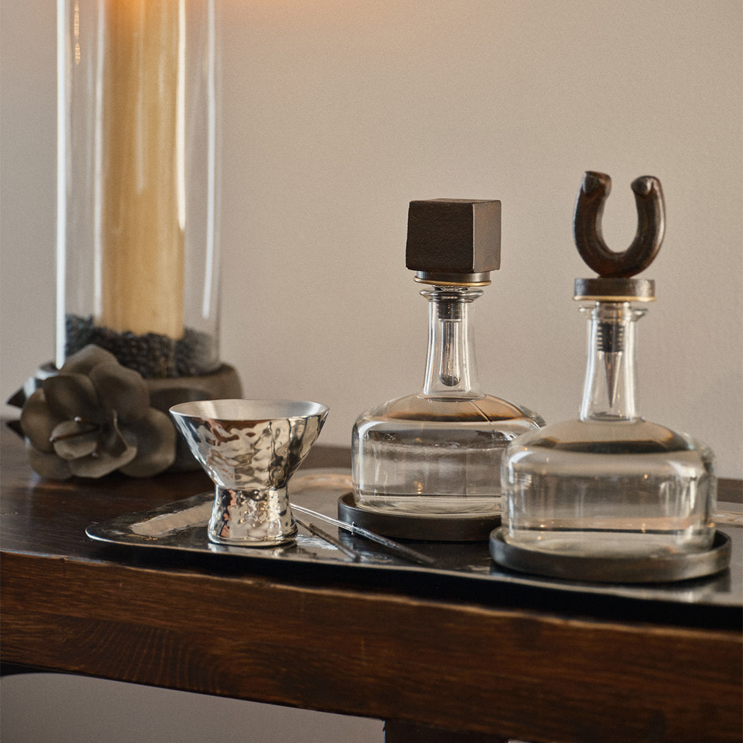 A wood table with a. silver tray and a iron rose with a glass and a xandle. On top of the tray is a silver bowl and two decanters with iron toppers.