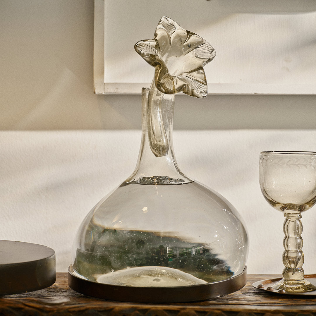 A clear mouth blown glass carafe on iron stand with glass flower cork.  Next to it id a clear glass cup. White wall as a background.