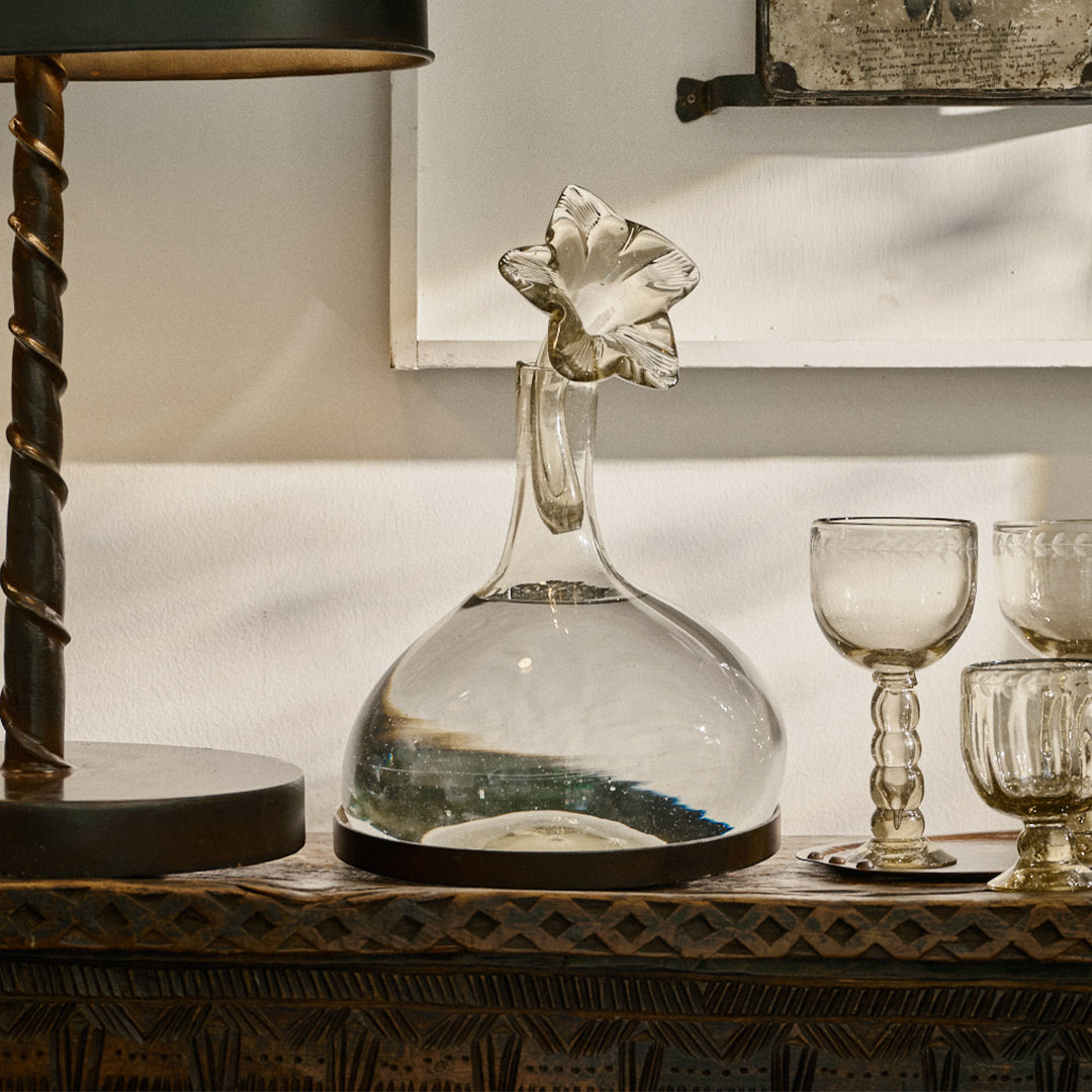 A lamp next to a clear mouth blown glass carafe on iron stand with glass flower cork and three glass cups.