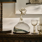 A lamp next to a clear mouth blown glass carafe on iron stand with glass flower cork and three glass cups.