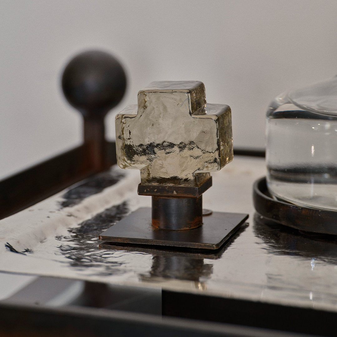 A mini mouth blown glass cross on iron stand. On top of a silver  object.  White background.