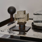 A mini mouth blown glass cross on iron stand. On top of a silver  object.  White background.