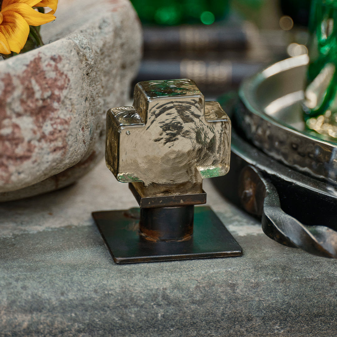 A mini mouth blown glass cross on iron stand on top of cement table. Outside background.