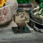 A mini mouth blown glass cross on iron stand on top of cement table. Outside background.