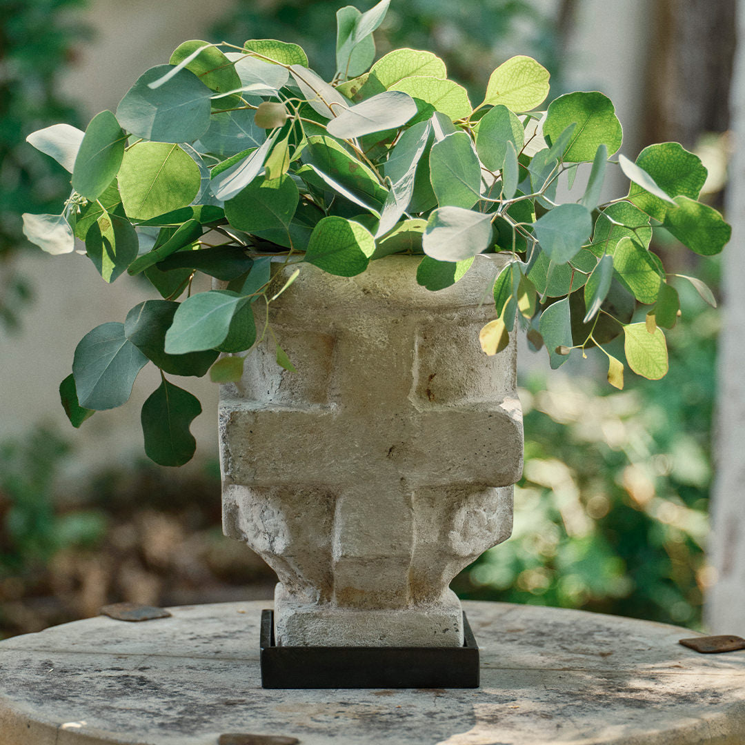 A stone container with a cross  on iron stand filled with green leaves on top of a table. 