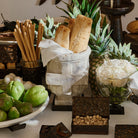 A white bowl filled with different green and white vegetables, some iron boxes  filled with nuts, a iron container with read sticks, a glass bowl with white roses, some pineapples and a  stone container with a cross  on iron stand filled with a white fabric and bread.