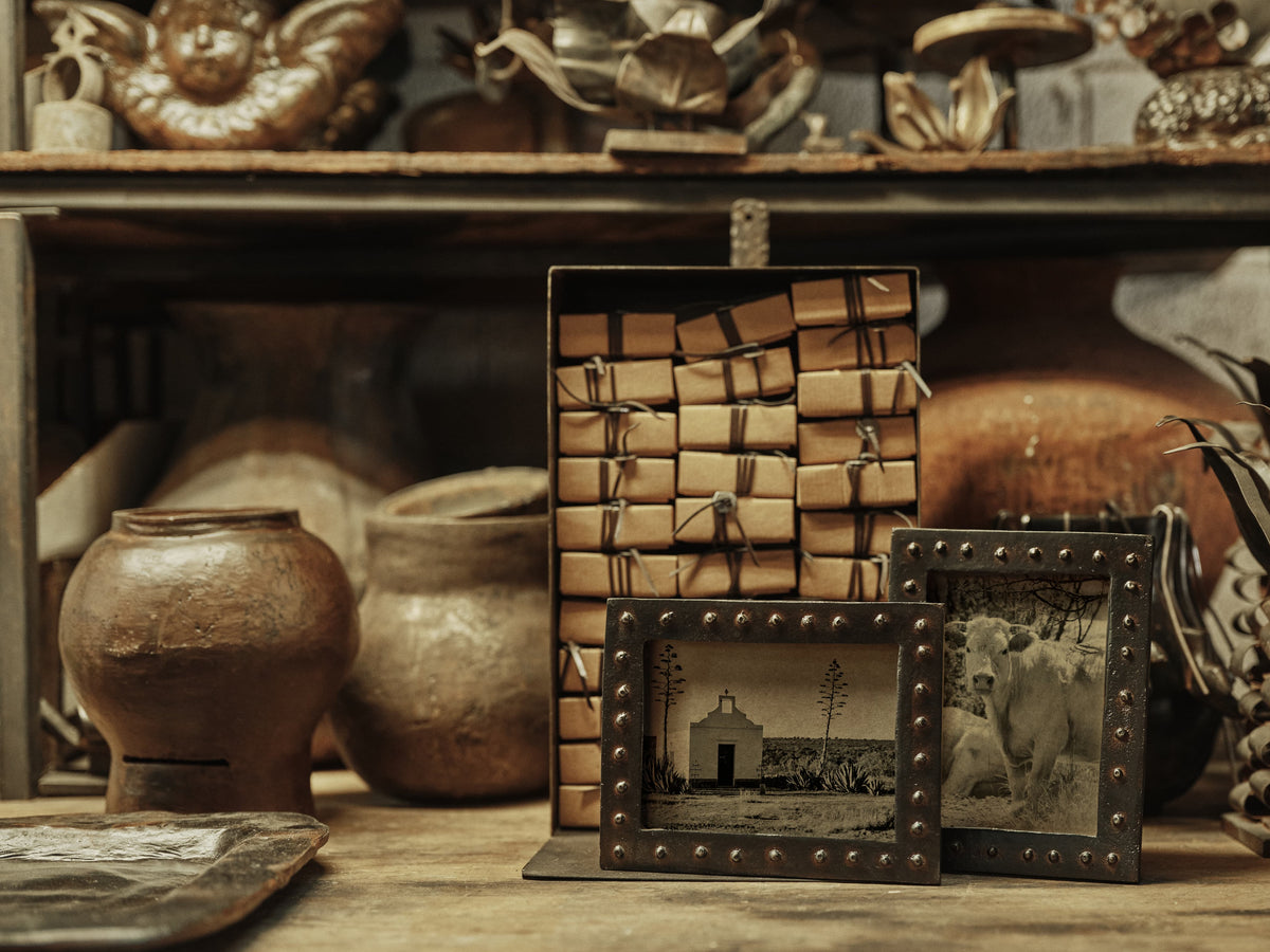 A styled product display featuring a stack of vintage leather-bound photo albums tied with string, flanked by two matching dark metal picture frames with brass studs, set against a rustic background of ceramic pottery and wooden shelves.