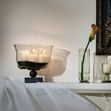 Three candles inside a fan-shaped vase resting atop a balin stand. On top of a white surface next to a flower inside a vase.