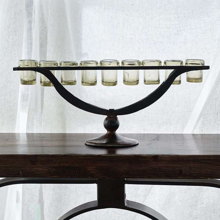 A nine votive holders supported by  forged iron outstretched arms supported by a wood table. On the background is a white fabric.