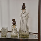 A mouth blown vibrant clear small angel decanter with iron angel halo topper next to a larger one  and two glasses on top of a tray.