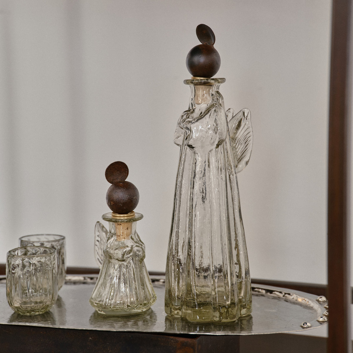 A mouth blown vibrant clear small angel decanter with iron angel halo topper next to a larger one  and two glasses on top of a tray.