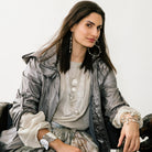 Woman seating wearing a greyish look. She is wearing several necklaces and bracelets. Also wearing hook earrings and looking at the front.