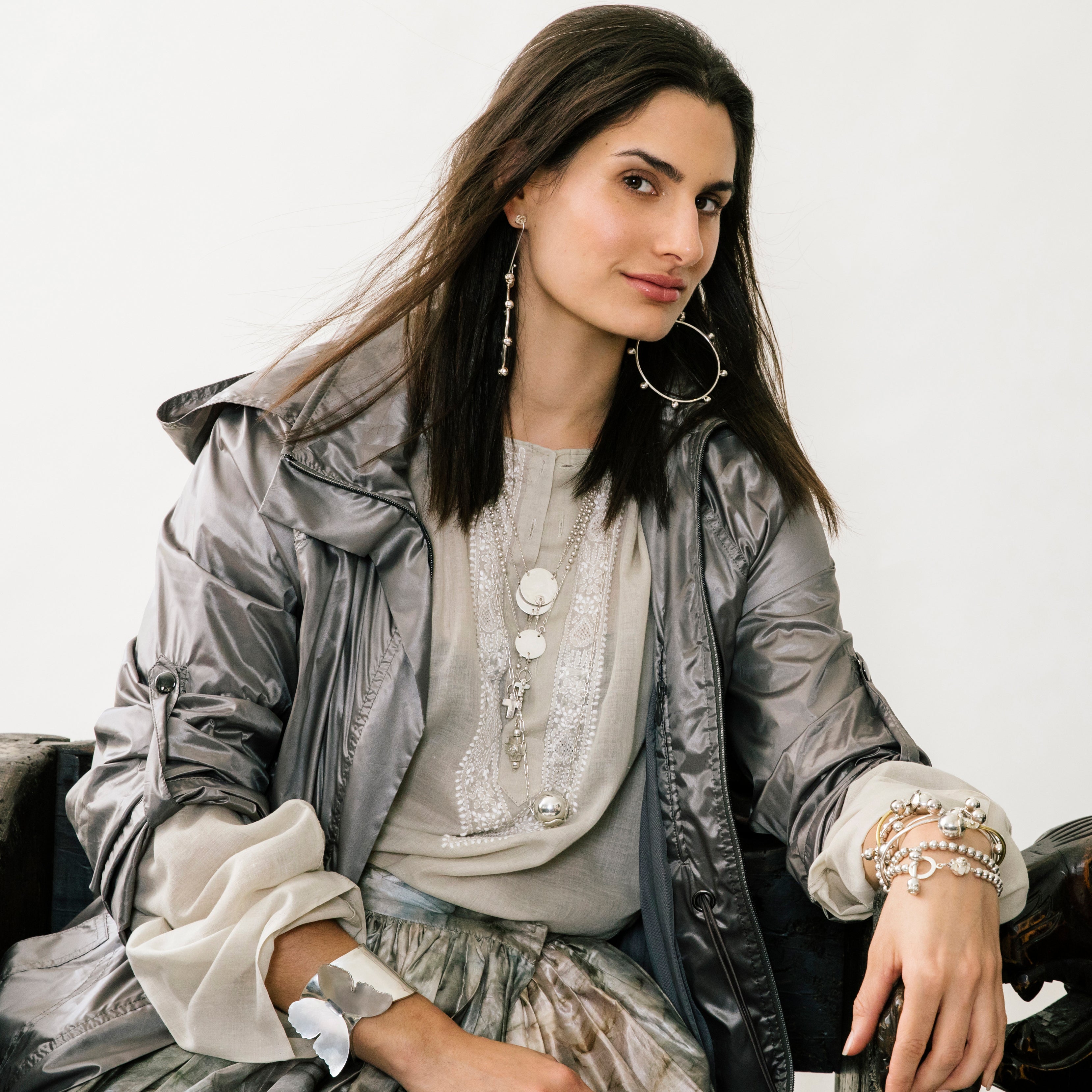 Woman seating wearing a greyish look. She is wearing several necklaces and bracelets. Also wearing hook earrings and looking at the front.