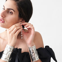 A brunette woman putting on a silver beads earring with her two hands while wearing silver cuffs. Plain background