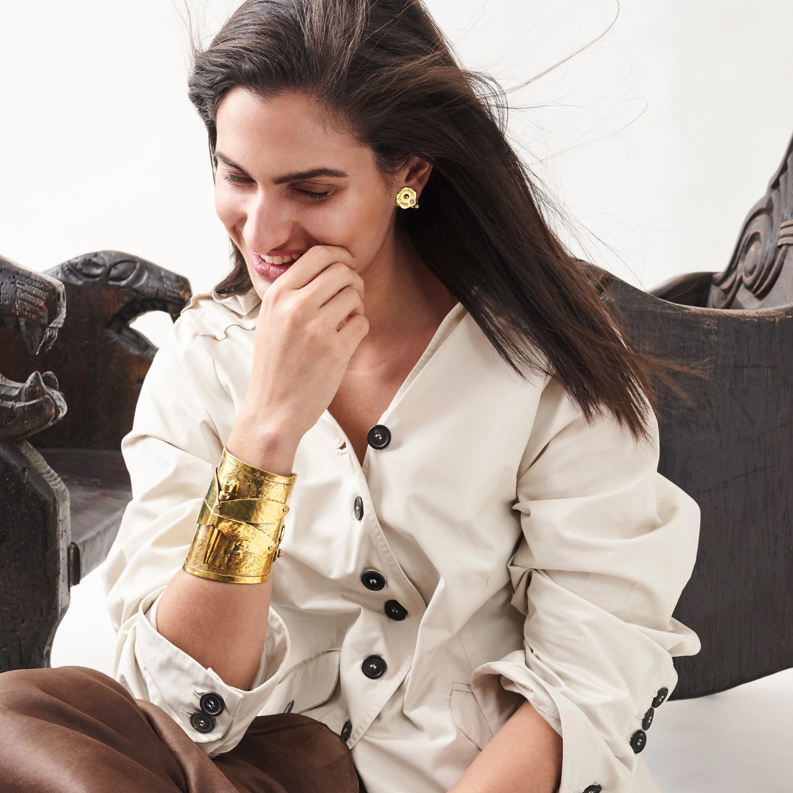 A women laughing with her  hand in her chin. Wind is blowing , she is seating on the floor resting her back on a dark bench. She is wearing gold earrings and a golden cuff.