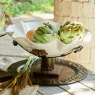 A white ceramic bowl with artichokes inside and a iron stand. On top of a iron  tray in a light colored wood surface. Outside background.