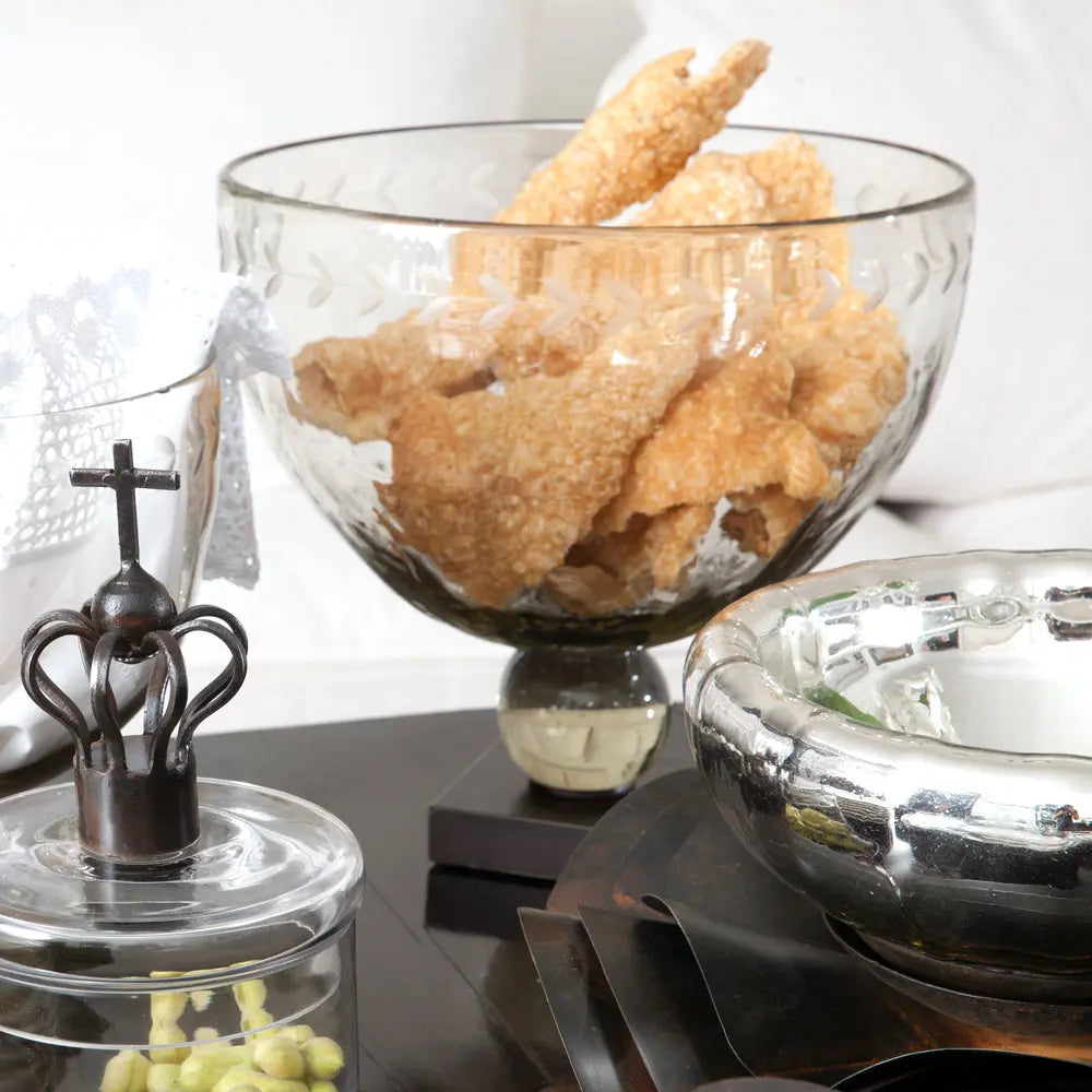 A mouth blown glass bowl with laurel pattern hand engraving on iron stand filled with food. Next to a bowl and a glass container.
