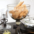 A mouth blown glass bowl with laurel pattern hand engraving on iron stand filled with food. Next to a bowl and a glass container.
