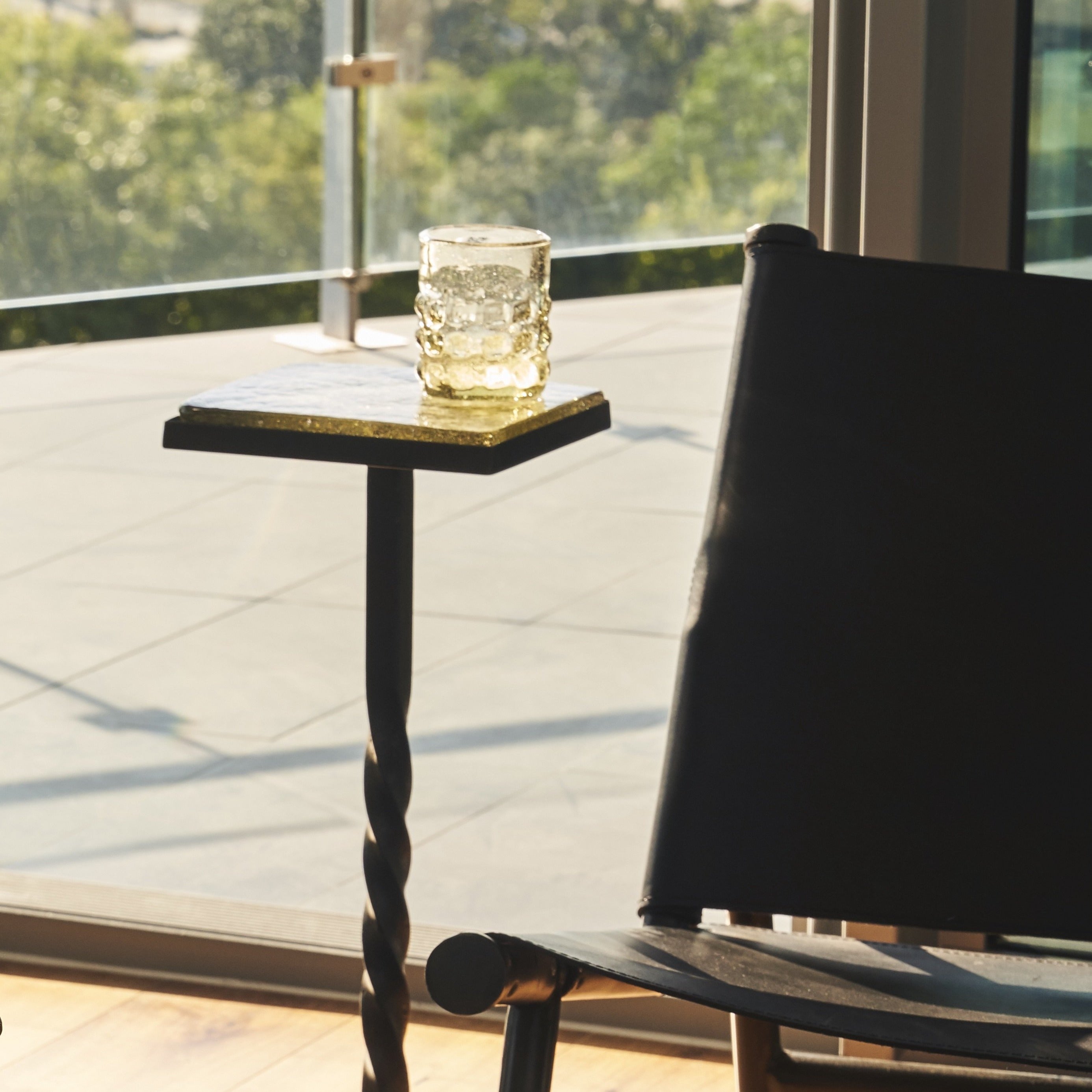 A glass on top of a small square table next to a dark chair. On the background is a window looking at a garden.