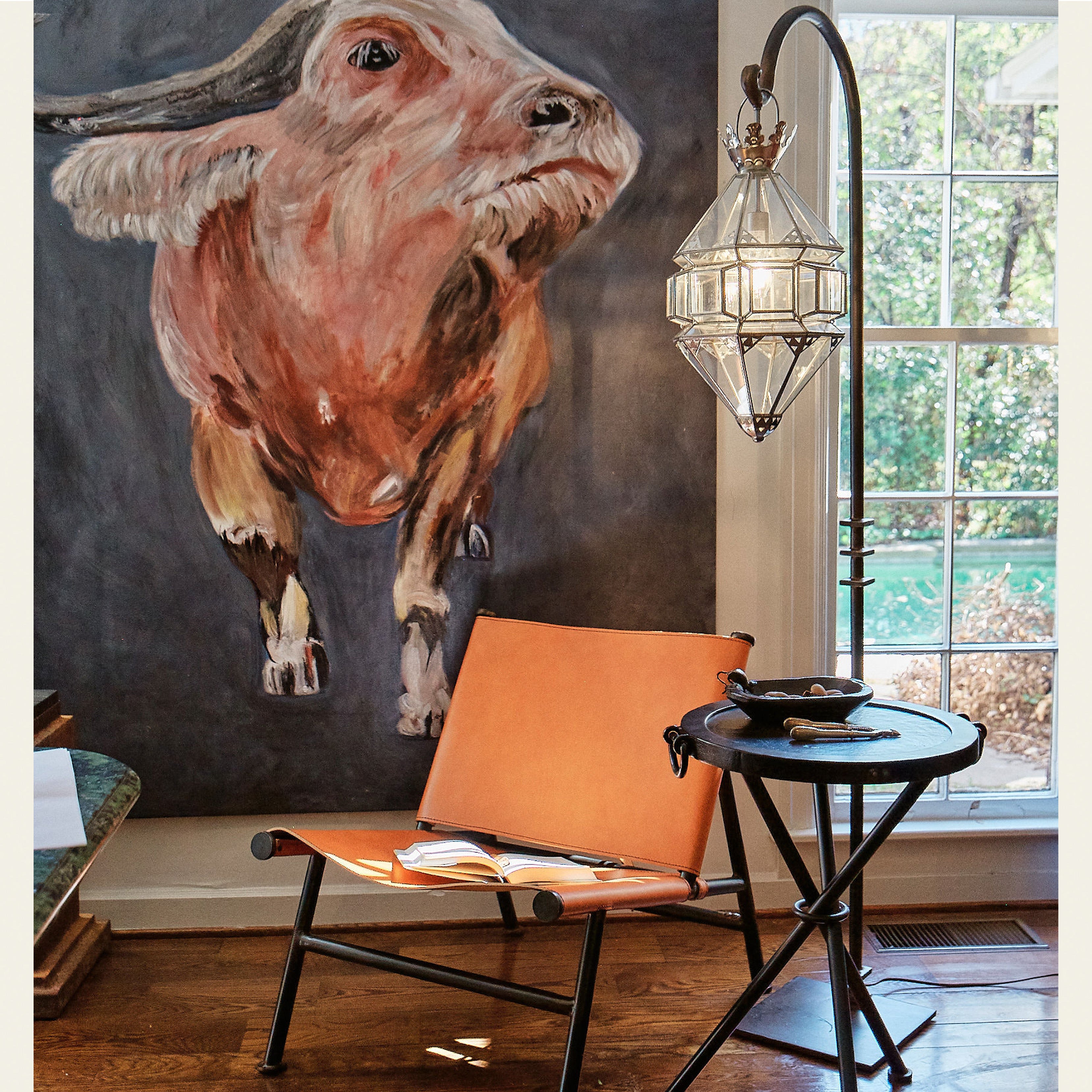 A light brown chair with a book on top. Next to a small round table with a ball and a  aluminum and glass lantern with hand-forged iron base.  Behind is a painting of a bull and a window.
