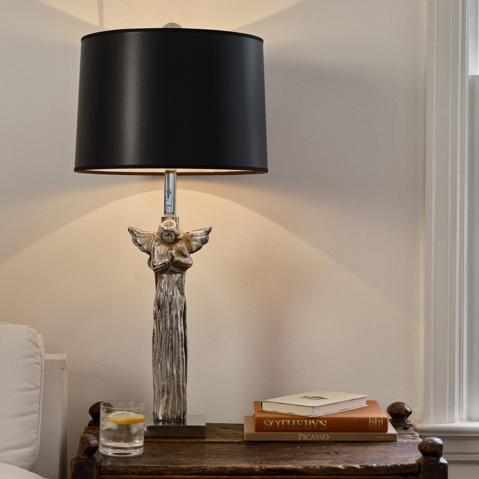 A wax cast nickel praying angels lamp. On top of a wooden table next to a glass of water and two books. Against a white wall.