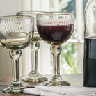 Three clear mouth blown goblets with  laurel engravings. One is filled with a deep red liquid. Next to them is a bottle with dark liquid. On the background is a window.