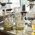 Three glasses, in one is a silver stick with a crown on it. Next to them is a decanter. In the background is a table with a dark bowl.