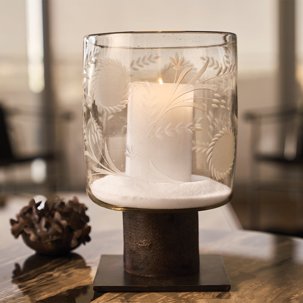 A glass vase with sunflower engravings  and a lit candle inside on top of an iron stand. Thee vase is on top of a wood surface.