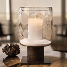 A glass vase with sunflower engravings  and a lit candle inside on top of an iron stand. Thee vase is on top of a wood surface.