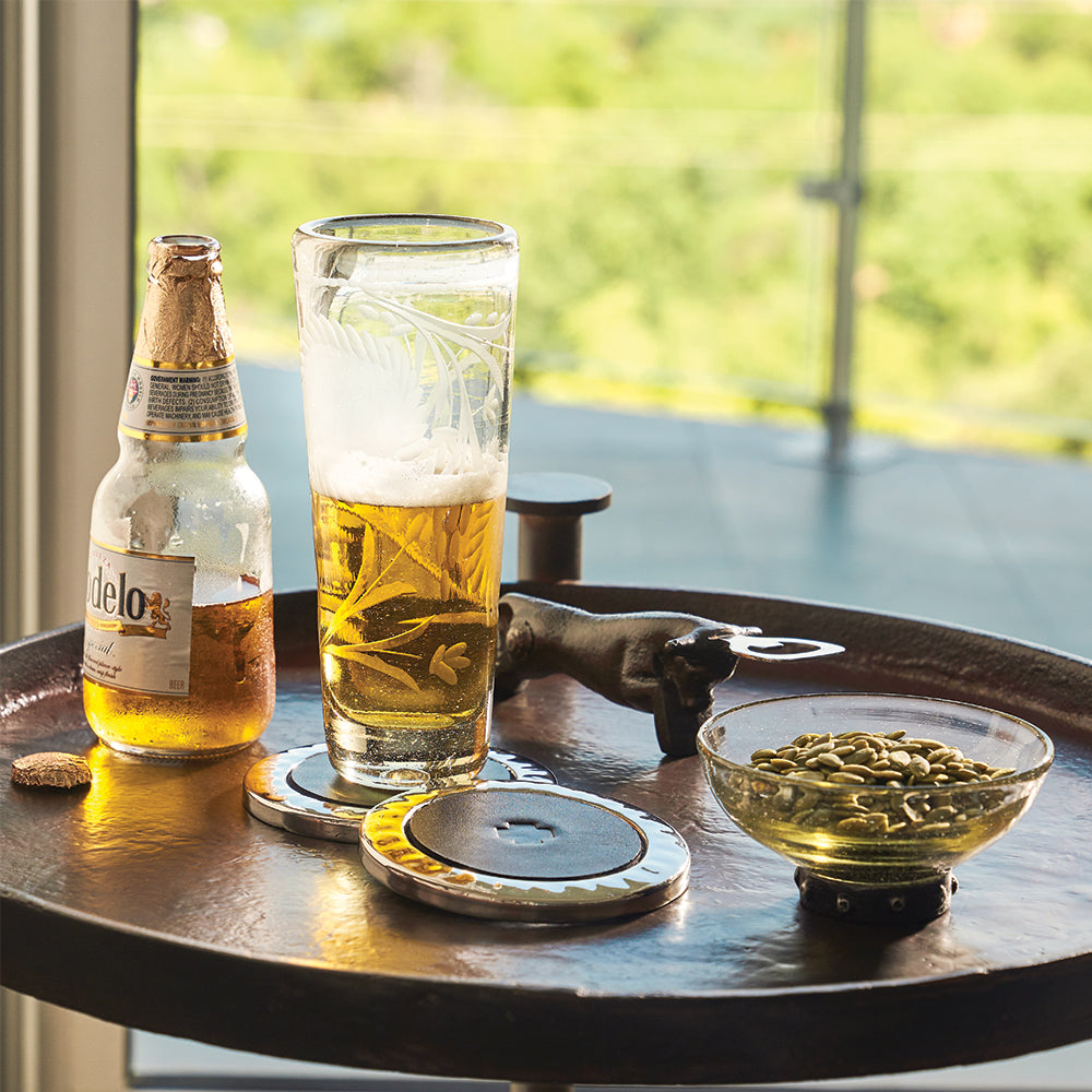 A beer bottle and a glass with beer next to a bowl with seeds and a bottle opener shaped as a bull. Everything on top of a table.