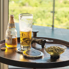 A beer bottle and a glass with beer next to a bowl with seeds and a bottle opener shaped as a bull. Everything on top of a table.