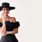 A woman smiling in a black dress and hat. In her arms are two silver cuffs.