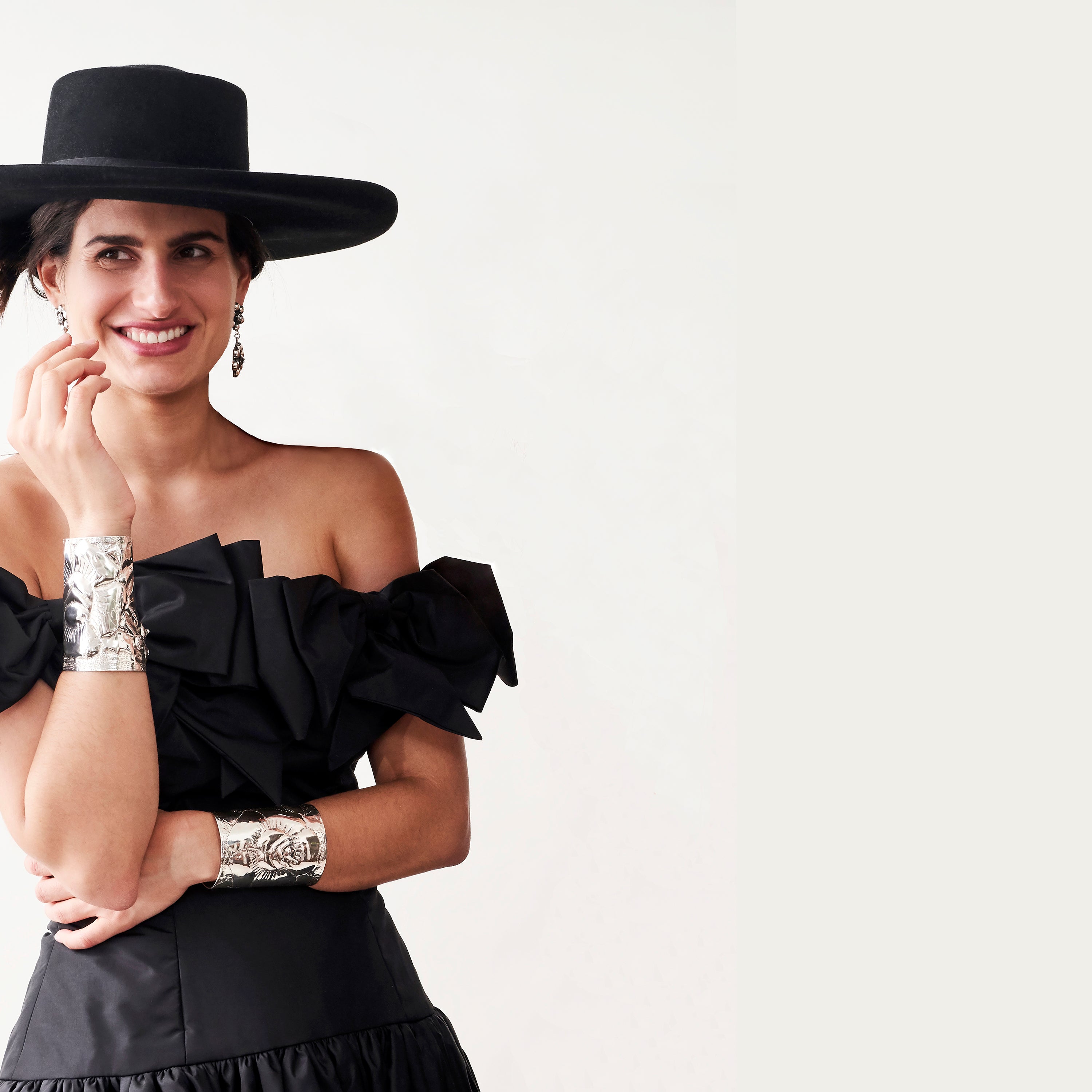 A woman smiling in a black dress and hat. In her arms are two silver cuffs.